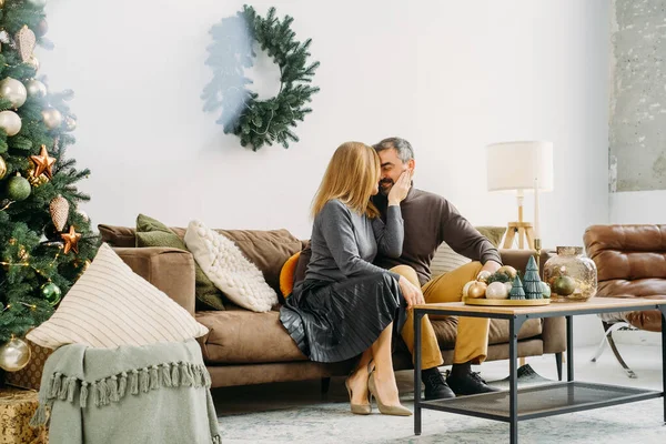 Couple celebrating romantic Christmas eve at home. Christmas interior decoration for family party. Copy space, greeting card. Middle-aged man and woman sitting on cozy couch, Tree and wreath