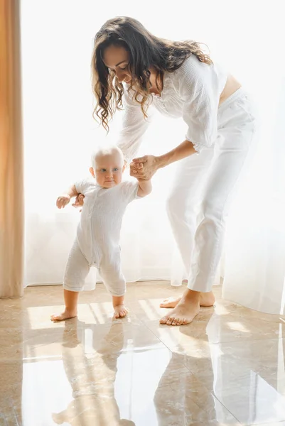 Mother helping to walk baby boy at home in a sunny room. Child learning to walk and keep balance with mothers support. First steps
