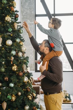Baba ve çocuk evde Noel ağacı süsler. Oğul babasının omuzlarında, uzun ağaca takılıyor..