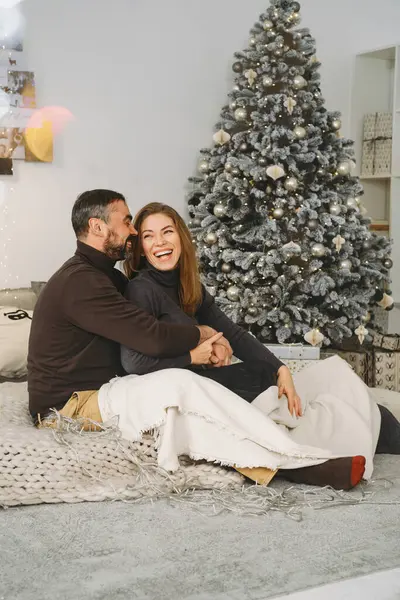 Happy couple celebrating romantic Christmas eve at home. Christmas interior decoration for family party. Copy space, greeting card. Middle-aged man and woman sitting on cozy bed with knitted blanket