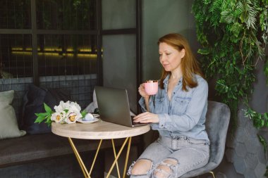 Kafede dizüstü bilgisayarda çalışan bir kadın. Orta yaşlı, kahve içip, yazı yazan ve okuyan bir serbest yazar. Çatı katında yeşil bitkiler var.