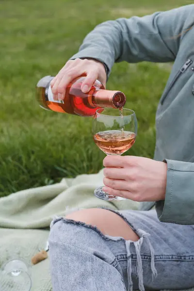 Woman uncork the bottle of rose wine on the picnic. Beautiful mature woman serve wine in glass outdoors on the grass. Woman at her 50s in the park