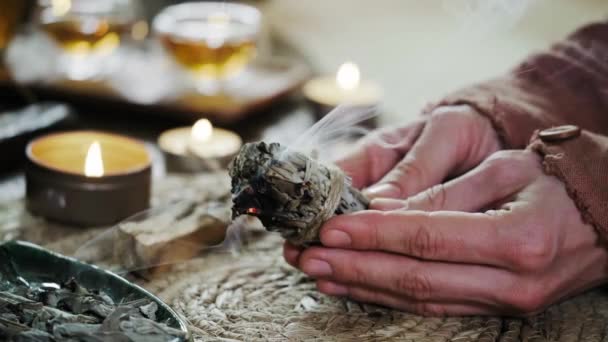 女人的手燃烧着白鼠狼 然后用蜡烛和绿色的植物在桌上做礼拜 烟熏治疗疼痛 明显的负面能量和冥想 — 图库视频影像