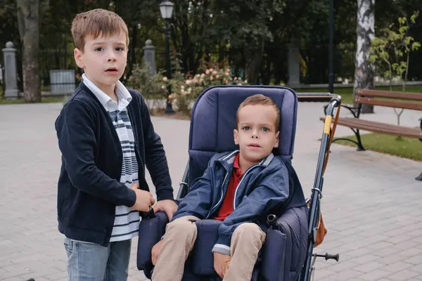 屋外を散歩する特別な車椅子に脳性麻痺を持つ子供 限られた能力を持つ人々の統合とアクセシビリティ 身体的な慢性的な健康状態の兄弟 — ストック写真