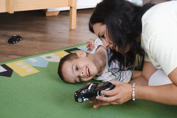 Disabled boy playing toy cars with mother at home. Cerebral palsy child entertaining on the mat with caregiver. Communication and rehabilitation in family