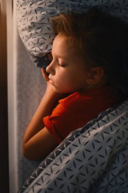Çocuk gece geç saatte yatakta uyuyor. Sıcak battaniyeyle örtülü şirin bebek kız yastığı. Çocuk gece dinleniyor. Işık komodine.