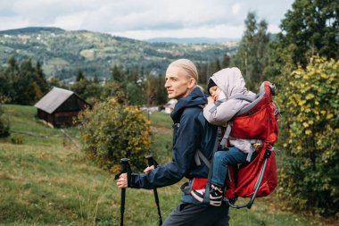 Sırt çantasında yorgun çocuğu olan bir gezgin. Babası kızını sırt çantasıyla taşıyor. Erkek çocuğu dağlara götürüyor. Çocuklarla seyahat etmek. Yavru bebekle sonbaharda yürüyüş, İskandinav yürüyüşü 