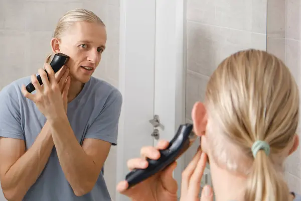 Man shaving whiskers hair by himself, self haircut at home in front of mirror in the bath. Male with long haircut using shaving machine trimmer. Electric razor for personal hygiene on quarantine
