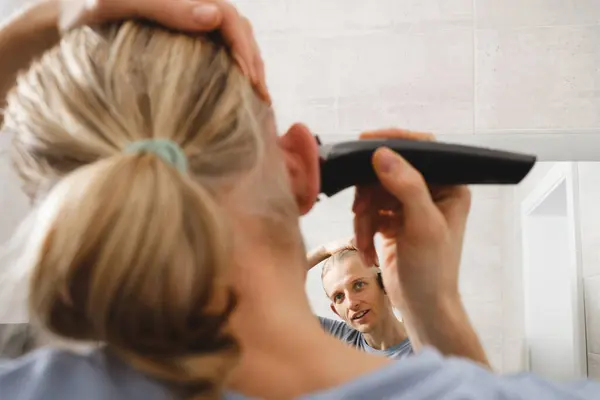 Man shaving whiskers hair by himself, self haircut at home in front of mirror in the bath. Male with long haircut using shaving machine trimmer. Electric razor for personal hygiene on quarantine