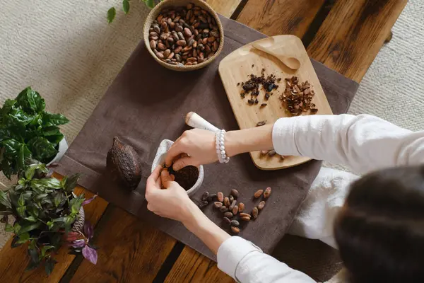 木のテーブルに有機カカオ豆を保持する女性の手 ココアニブ 儀式のための素朴なブーフスタイルで作る職人チョコレート チョコレート作り ポンドクローズアップトップビュー — ストック写真