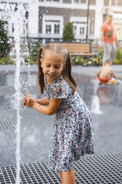 Şehirdeki çeşmede oynayan bir çocuk. Sıcak yaz havası. Su fıskiyeleriyle oynayan, koşan ve gülen mutlu kız. Aktif çocukluk. Açık havada oyunlar. Modern şehir sokağı