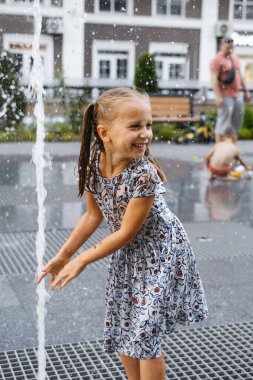 Şehirdeki çeşmede oynayan bir çocuk. Sıcak yaz havası. Su fıskiyeleriyle oynayan, koşan ve gülen mutlu kız. Aktif çocukluk. Açık havada oyunlar. Modern şehir sokağı