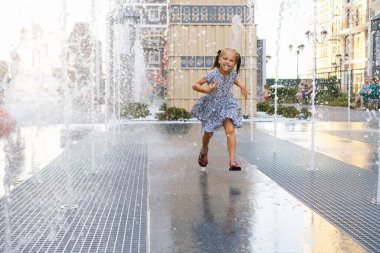 Sıcak yaz havası. Şehirdeki çeşmede oynayan bir çocuk. Su fıskiyeleriyle oynayan, koşan ve gülen mutlu kız. Aktif çocukluk. Açık havada oyunlar. Modern şehir sokağı