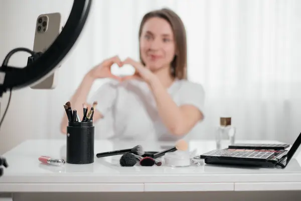 Blogger showing cosmetics and heart shape Woman doing make-up at home, apply decorative cosmetics. Beautiful young female using brush near the mirror and smartphone.