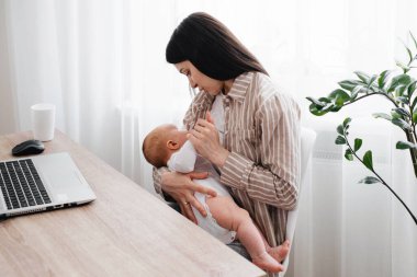 Evde, pencerenin üzerinde dizüstü bilgisayarının yanında bebeği olan, çalışan ve emziren bir kadın. Bebekle bilgisayarın yanında annelik eğitimi alıyorum. Çocuklu bir anne olarak evde çalışıyorum.