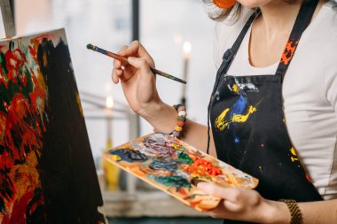 Yağlı boya boya ve fırçalarla gerçek bir stüdyoda çalışan ressam tuval ve modern sanat eserleri yaratıyor. Pencerenin oradaki genç bayan, yakın plan.