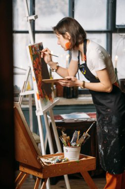 Yağlı boya boya ve fırçalarla gerçek bir stüdyoda çalışan ressam tuval ve modern sanat eserleri yaratıyor. Pencerenin oradaki genç bayan, yakın plan.