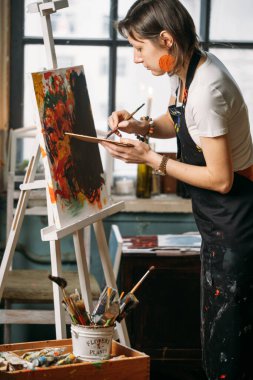 Yağlı boya boya ve fırçalarla gerçek bir stüdyoda çalışan ressam tuval ve modern sanat eserleri yaratıyor. Atölyenin penceresindeki genç kadın.