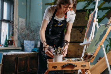 Yağlı boya boya ve fırçalarla gerçek bir stüdyoda çalışan ressam tuval ve modern sanat eserleri yaratıyor. Penceredeki genç kadın, atölye anı