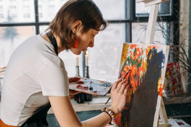 Yağlı boya boya ve fırçalarla gerçek bir stüdyoda çalışan ressam tuval ve modern sanat eserleri yaratıyor. Pencerenin oradaki genç bayan, yakın plan.