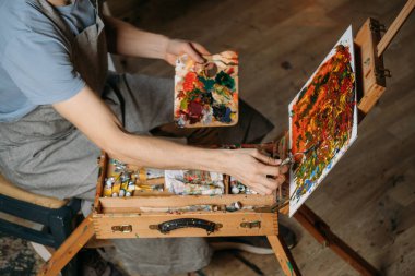 Yağlı boya boya ve fırçalarla gerçek bir stüdyoda çalışan ressam tuval ve modern sanat eserleri yaratıyor. Paletli genç adam, yakın plan.
