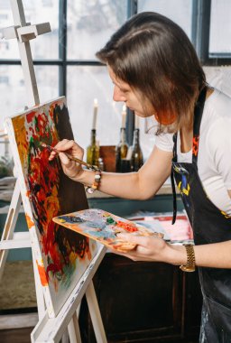 Yağlı boya boya ve fırçalarla gerçek bir stüdyoda çalışan ressam tuval ve modern sanat eserleri yaratıyor. Pencerenin oradaki genç bayan, yakın plan.