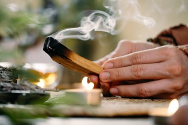 Kadın elleri Palo Santo 'yu yakmadan önce, masada mumlar ve yeşil bitkilerle. Leke dumanı acıyı ve stresi tedavi eder. Negatif enerjiyi temizler. Meditasyon tahtası.
