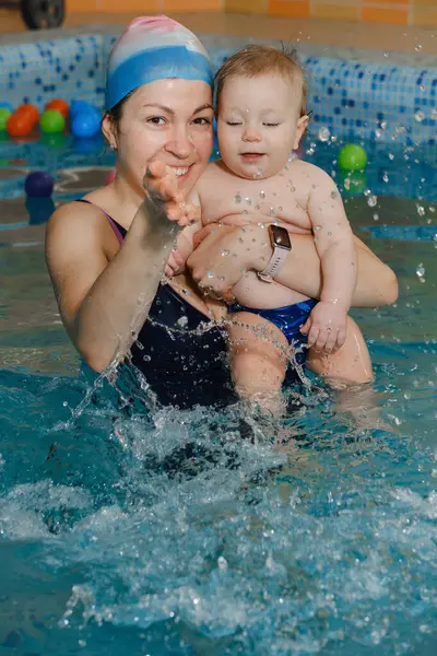 stock image Early swimming coach training to swim baby boy in indoor pool. Playing activity for infant with mother, child physical development in water with joy and fun. Accessories and toys for diving lesson