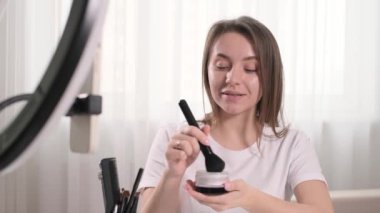 Kadın blogcu evde makyaj yapıyor, dekoratif makyaj yapıyor, pudra sürüyor. Oturma odasındaki aynanın yanında fırça kullanan güzel genç bir kadın, kullanıcılar için kişisel bakım ve ürün sunumu İnternet 'te 