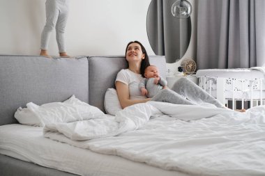 Annem ve iki çocuğum evde, yatak odasında. Bebek annesinin ellerinde dinlenirken, abla oyun oynarken, hareket ederken, yatağın yanında zıplarken, küçük kızıyla konuşurken bir bebeğe sarılırken,