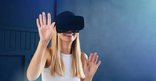 Sarı saçlı genç bir kadın sanal ortamla iletişim kurarken VR kulaklık takıyor ve gülümsüyor..