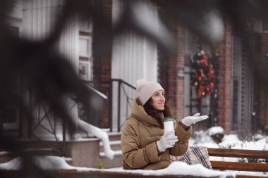 Neşeli esmer kadın kışın şehirde açık hava matarasından su içiyor. Isınmak, anın tadını çıkarmak, karla kaplı ağaçların üzerinde fren yapmak sıcak içecekle yeniden kullanılabilir başlık. Bankta oturan kadın.