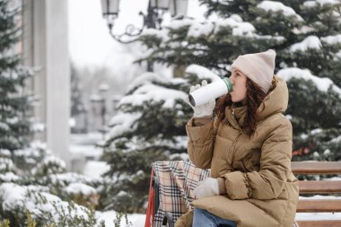 Neşeli esmer kadın kışın şehirde açık hava matarasından su içiyor. Isınmak, anın tadını çıkarmak, karla kaplı ağaçların üzerinde fren yapmak sıcak içecekle yeniden kullanılabilir başlık. Bankta oturan kadın.