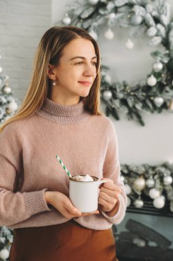 Elinde şekerlemelerle kakao tutan kazaklı bir kadın. Noel arifesinde evde lezzetli sıcak çikolata kaplı tatil atmosferiyle. Konforlu Noel arkaplanı, çözülmüş ağaç, ışıklar.