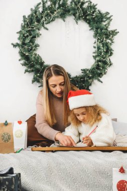 Yeni yürümeye başlayan çocuk annesiyle Noel 'i bekliyor. Şirin kıvırcık kız için şenlik havası. Çocuk İskandinav iç mekanında Noel ağacının yanına Noel şapkasını koyuyor.
