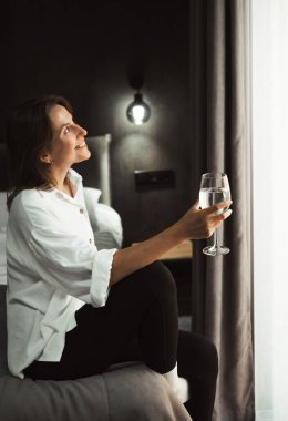 Kadın rahat bir otel yatağının kenarında oturur, pencereden dışarı bakar ve bir bardak suyla hayal kurar. Seyahat tecrübelerini ve planlarını yansıtan kişi, bavullarıyla çevrili..