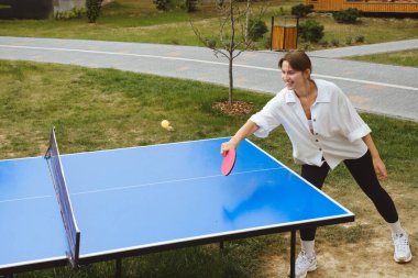 Kadın dışarıda masa başında pinpon oynuyor, topa odaklanıyor. Yaz tatillerinde açık hava aktiviteleri ve dostça bir rekabet