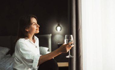 Kadın rahat bir otel yatağının kenarında oturur, pencereden dışarı bakar ve bir bardak suyla hayal kurar. Seyahat tecrübelerini ve planlarını yansıtan kişi, bavullarıyla çevrili..