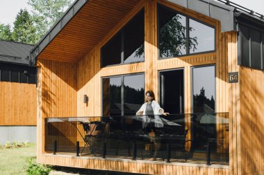 Modern bir otel odasında dinlenen bir kadın. Ahşap evin balkonunda duran bir kadın. Yaz tatili ve açık havada seyahat. Dinlenme için rahat bir ev.