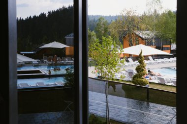 Pencereden yemyeşil manzaralı modern bir otel havuzunda huzurlu bir günün tadını çıkar. Güneşli bir öğleden sonra insanlar ferahlatıcı suda gevşeyip dinlenirken güneşlenirler..