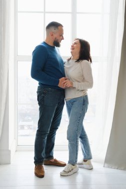 40 'lı yaşlarda sevgili bir çift büyük pencerenin önünde birlikte güzel vakit geçirmenin tadını çıkarıyor. Aile gülümsemesi, sarılma, sevinç anı ve birliktelik. 