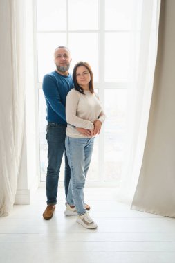 40 'lı yaşlarda sevgili bir çift büyük pencerenin önünde birlikte güzel vakit geçirmenin tadını çıkarıyor. Aile gülümsemesi, sarılma, sevinç anı ve birliktelik. 