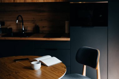 Ahşap masa, açık bir kitap ve mutfakta beyaz bir fincan kahve. Rahatlama ve rahat anlar için mükemmel bir yer.