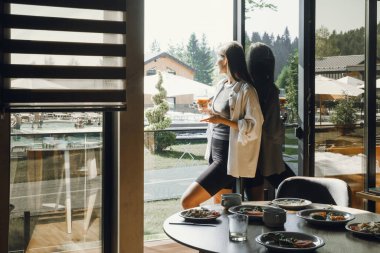 Manzaralı modern bir otel yemek salonunda dinleniyor. Kadın gezgin, çağdaş bir otelin geniş penceresinden dışarı bakarken içki içmekten zevk alır. Masa lezzetli yemekler ve rahat bir atmosferle dolu.