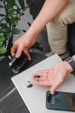 Modern dijital işitme cihazları, akıllı telefon ve şarj çantası şık bir ofis masasında, iş için hazırlanmış cihazlar. İş ortamı, ulaşılabilirlik ve profesyonel yaşam tarzı. Adam kulaklık kullanıyor. 