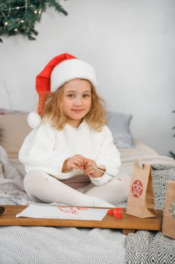 Noel Baba şapkalı küçük bir kız Noel mektubu yazar ve güzel bir tatil odasına kırmızı boya çizer. Şenlikli ambalaj ve dekorla..