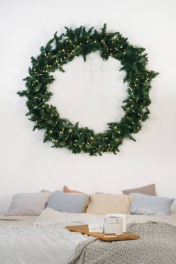 Noel için dekore edilmiş minimalist yatak odası büyük yeşil bir çelenk, peri ışıkları, rahat yastıklar ve yatağın üzerinde tatil hediyeleri..