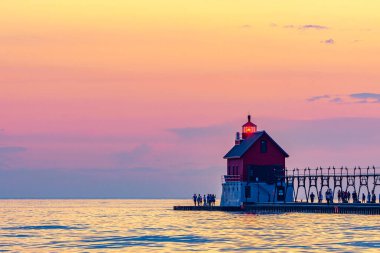 Bir grup insan Grand Haven, Michigan, deniz feneri ve iskelede sıcak, pastel bir gün batımı izliyor. Yüksek kalite fotoğraf