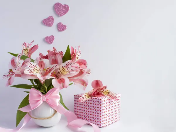 一束粉红色的花 装在一个白色的小花瓶里 旁边有一个礼物 上面有一朵花 上面有小粉色的心 你可以看到四个粉色的心 在白色的背景上 — 图库照片