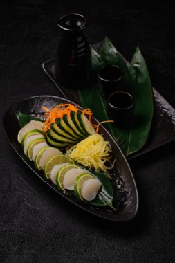 Scallop sashimi with vegetables and a bottle of sake on black concrete background